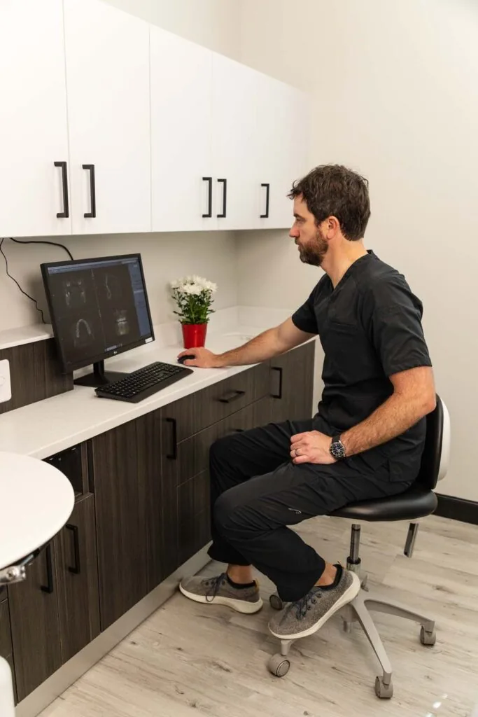 Dr. Holmes in dark scrubs seated on a stool, reviewing MRI or CT scans on a computer in a clean clinical room. The desk includes upper and lower cabinets, a small potted plant, and a monitor displaying medical imaging. The space features white walls and light flooring.