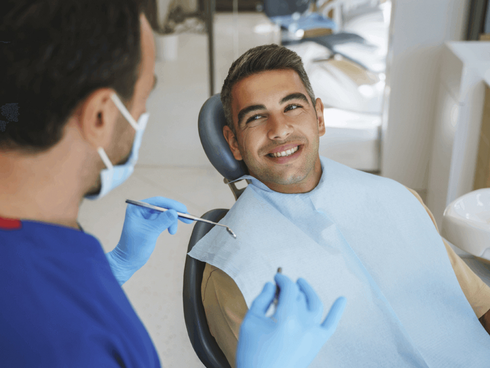 patient wearing a dental bib sitting in exam chair smiling as dentist gets ready check teeth