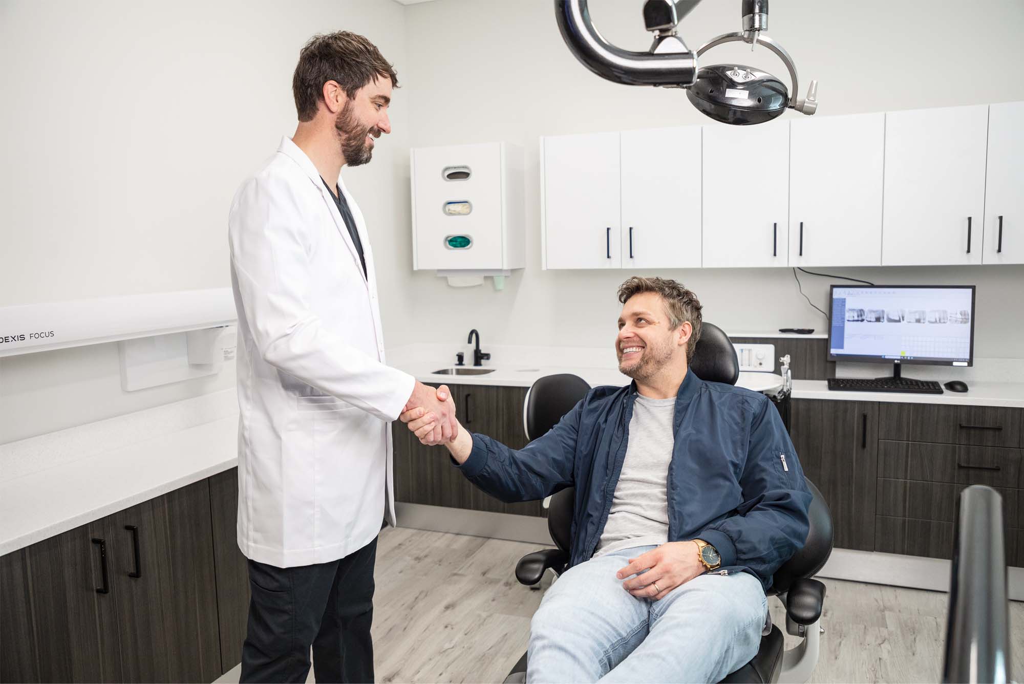 Dr. Holmes in a white coat shaking hands with a seated patient wearing a blue jacket and light blue jeans in a modern dental operatory. The room includes a dental chair, overhead light, computer monitor displaying dental images, and cabinetry with a sink. The interaction appears friendly, suggesting the end of a consultation or procedure.