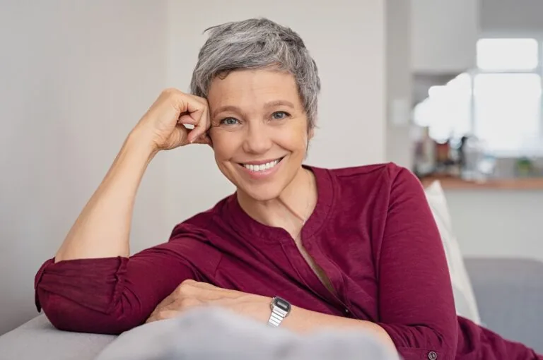 Smiling older adult with short gray hair wearing a maroon shirt, seated comfortably in a home setting—showcasing the natural look and confidence provided by hybrid dentures.