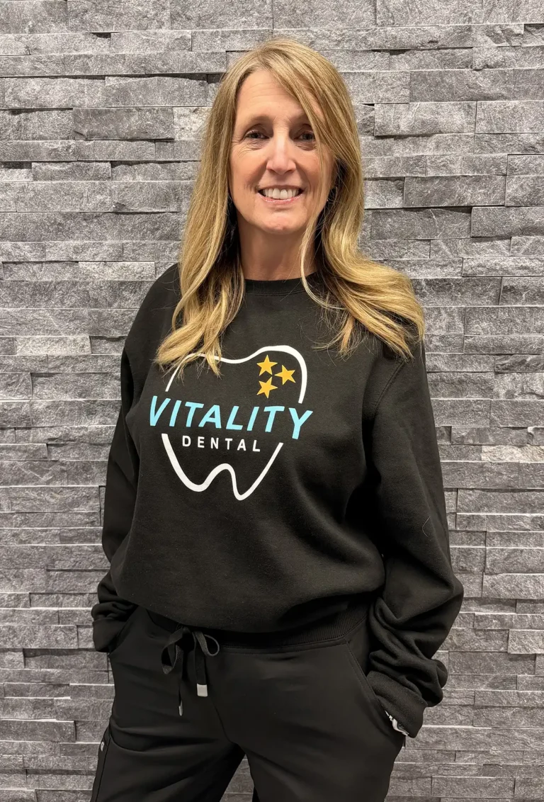 Renae hygiene assistant at Vitality Dental smiling in front of a textured wall. She provides friendly and supportive care to help patients feel comfortable during their visits.