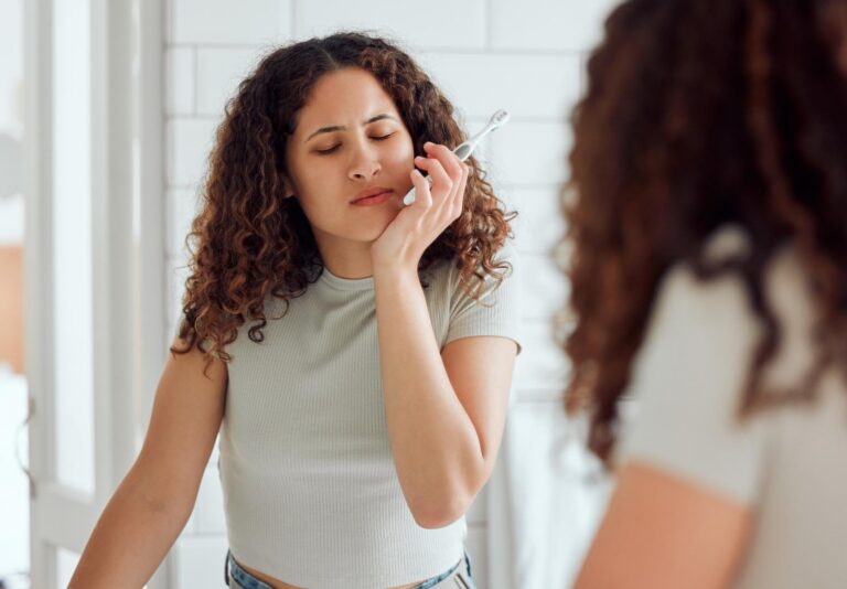 Woman with curly hair holding toothbrush and touching cheek in pain, standing in bathroom mirror, showing signs of toothache or dental emergency.